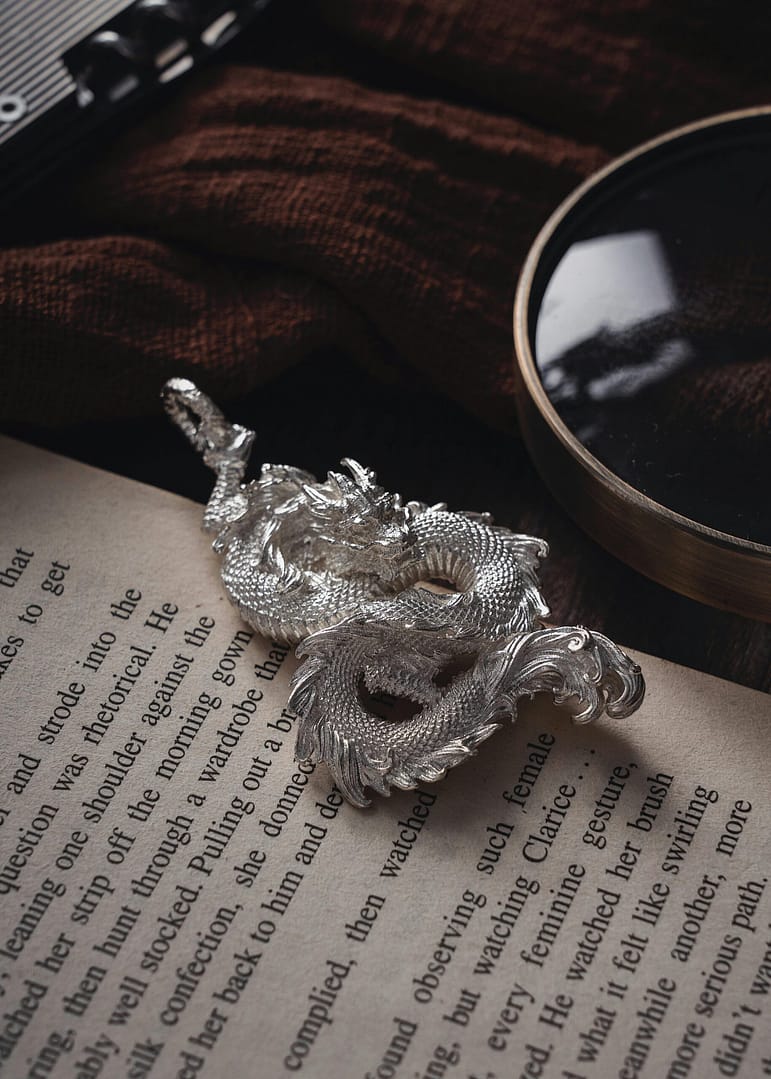 Artistic silver dragon placed on vintage book with magnifying glass detail.